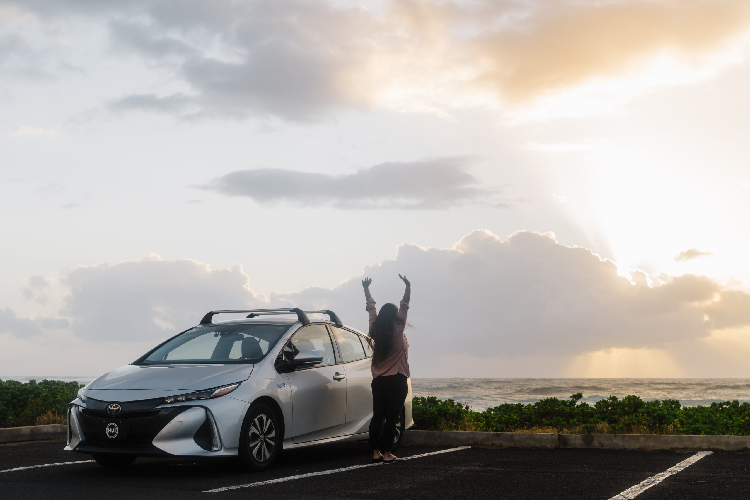 Traveler stretching in the early morning next to a Hui Car Share vehicle at Sandy Beach enjoying Oahu on a budget